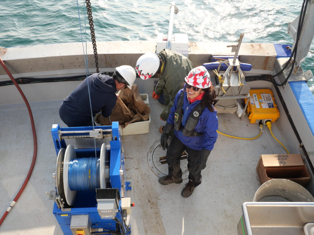 A crew of three people on the deck of a boat. Women with helmet on looking up and smiling